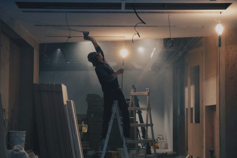 A person fire sealing the ceiling while standing on a ladder in a dimly lit room.