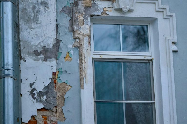 Peeling paint and damaged plaster on a wall next to a window.