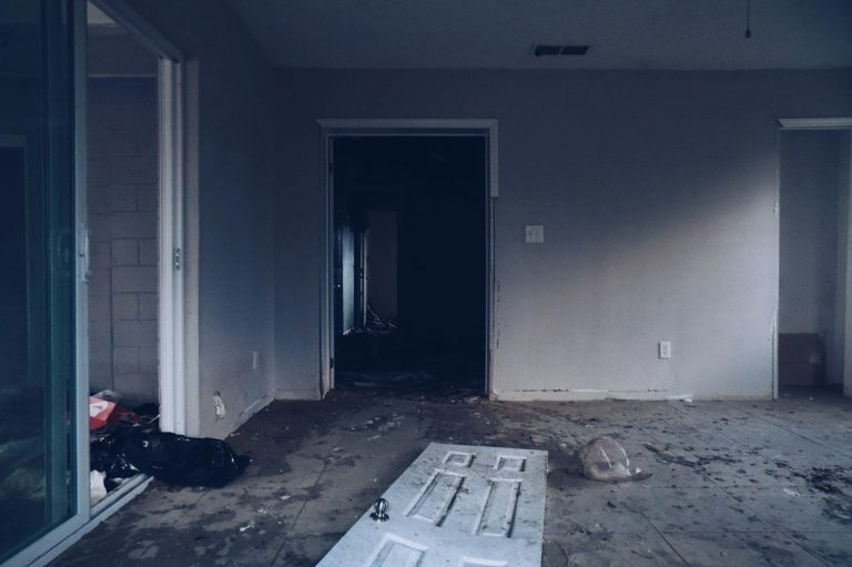 Empty, partially furnished room with grey walls and a dirty floor. Light coming from a window.